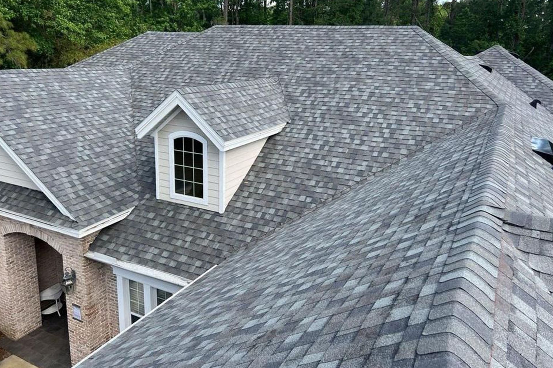 Gray shingle roof on a house with a white dormer and window. Trees are in the background.