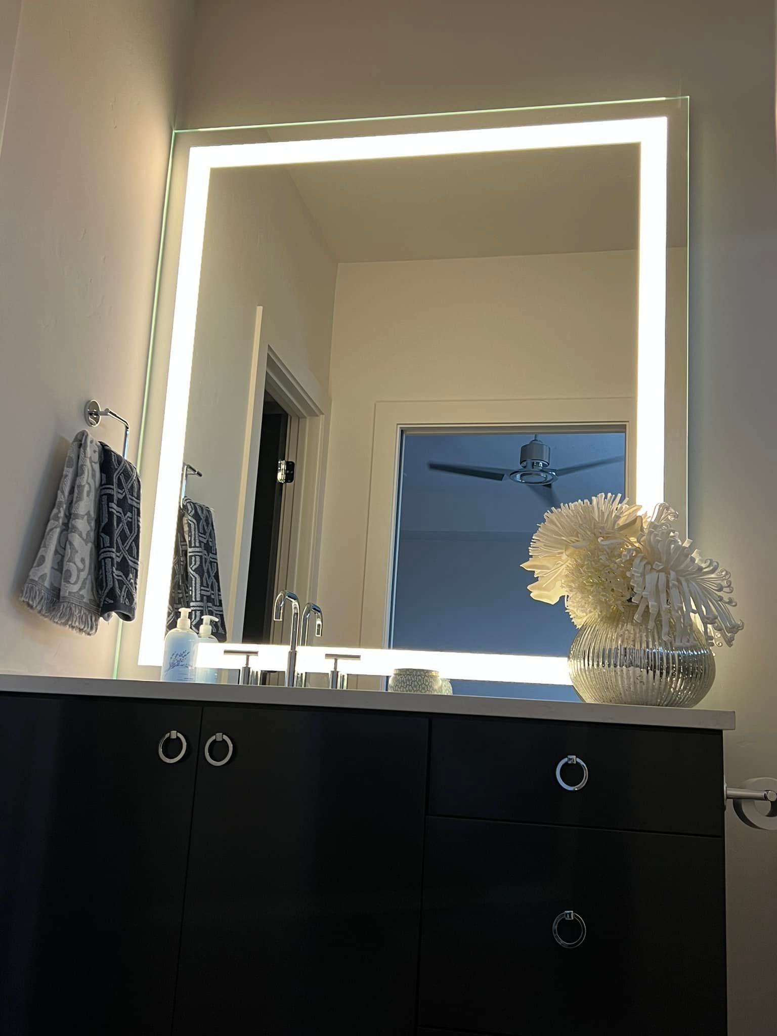 Lit rectangular mirror above a dark cabinet in a bathroom; white flowers and towels are visible.
