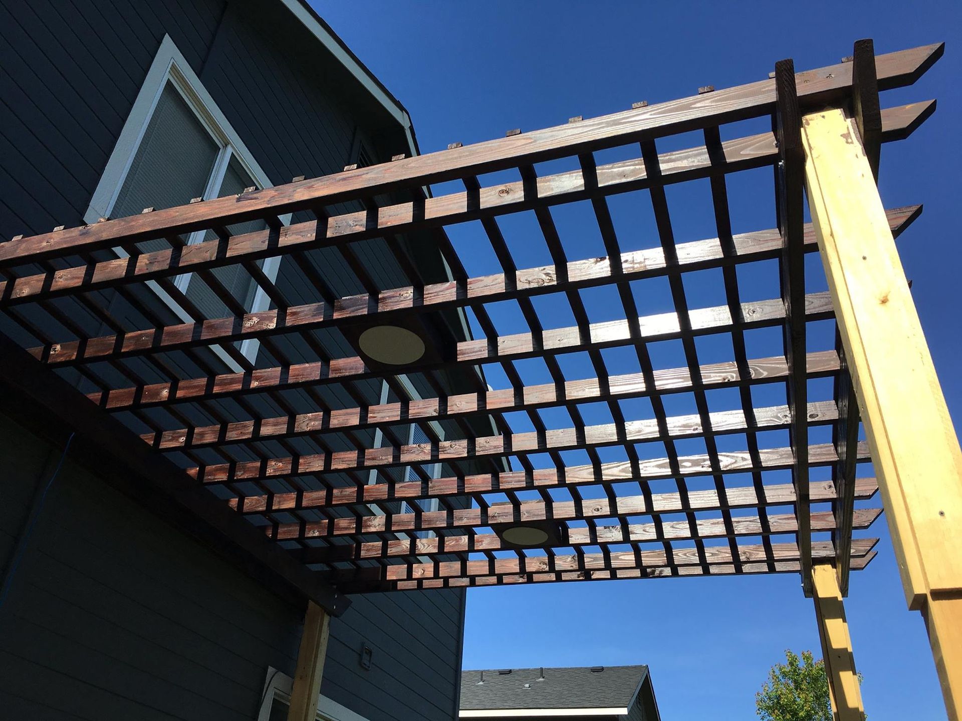 Brown wooden pergola with grid pattern against a blue sky, attached to a gray house.