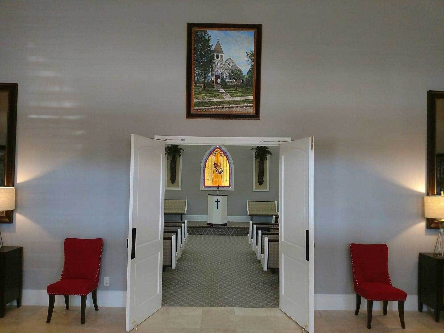 Interior of a chapel, viewed from open double doors. Red chairs, art, and stained glass are visible.