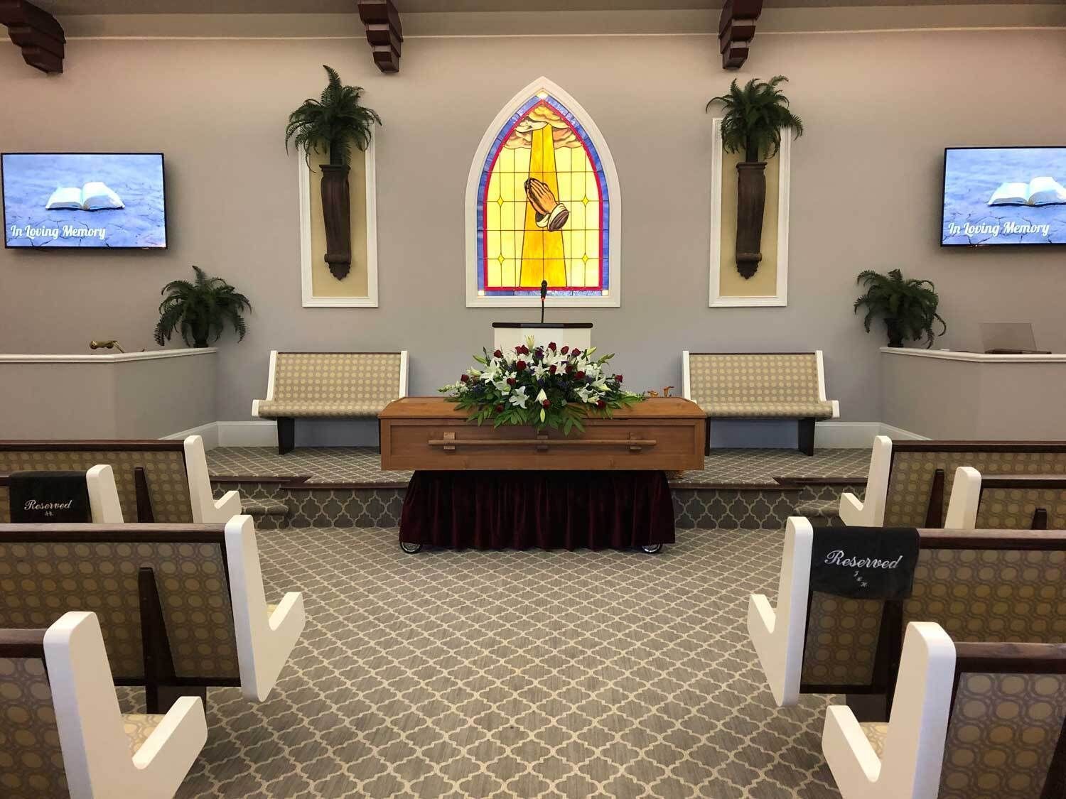 Interior of a funeral chapel with a casket, floral arrangement, and rows of chairs.