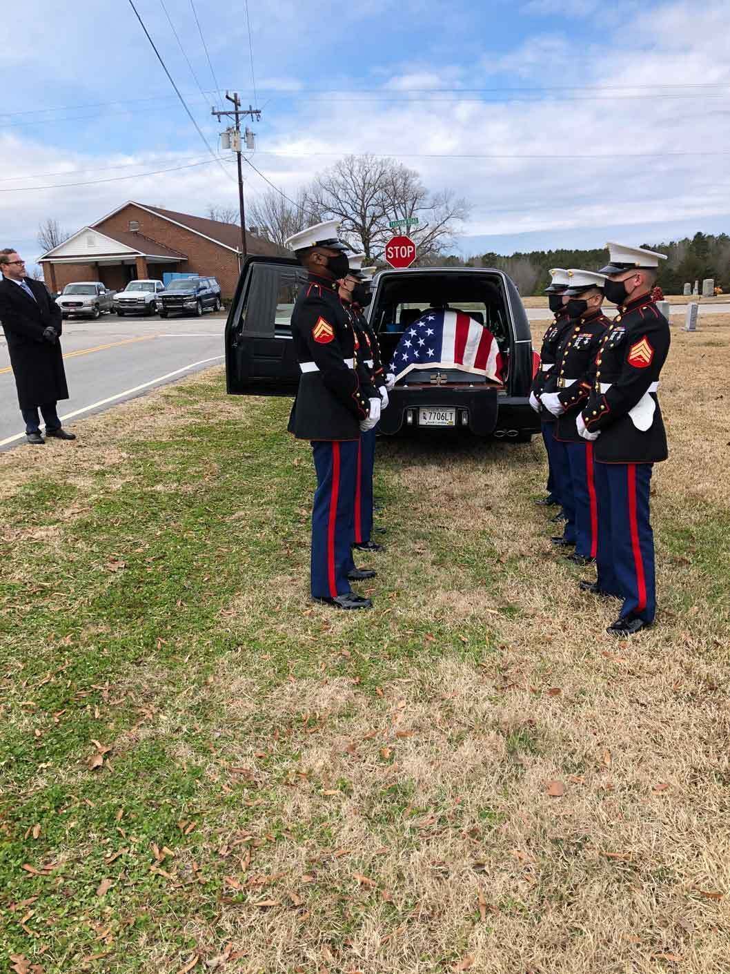 Marines in dress blues stand at attention beside a hearse with an American flag draped over the casket.