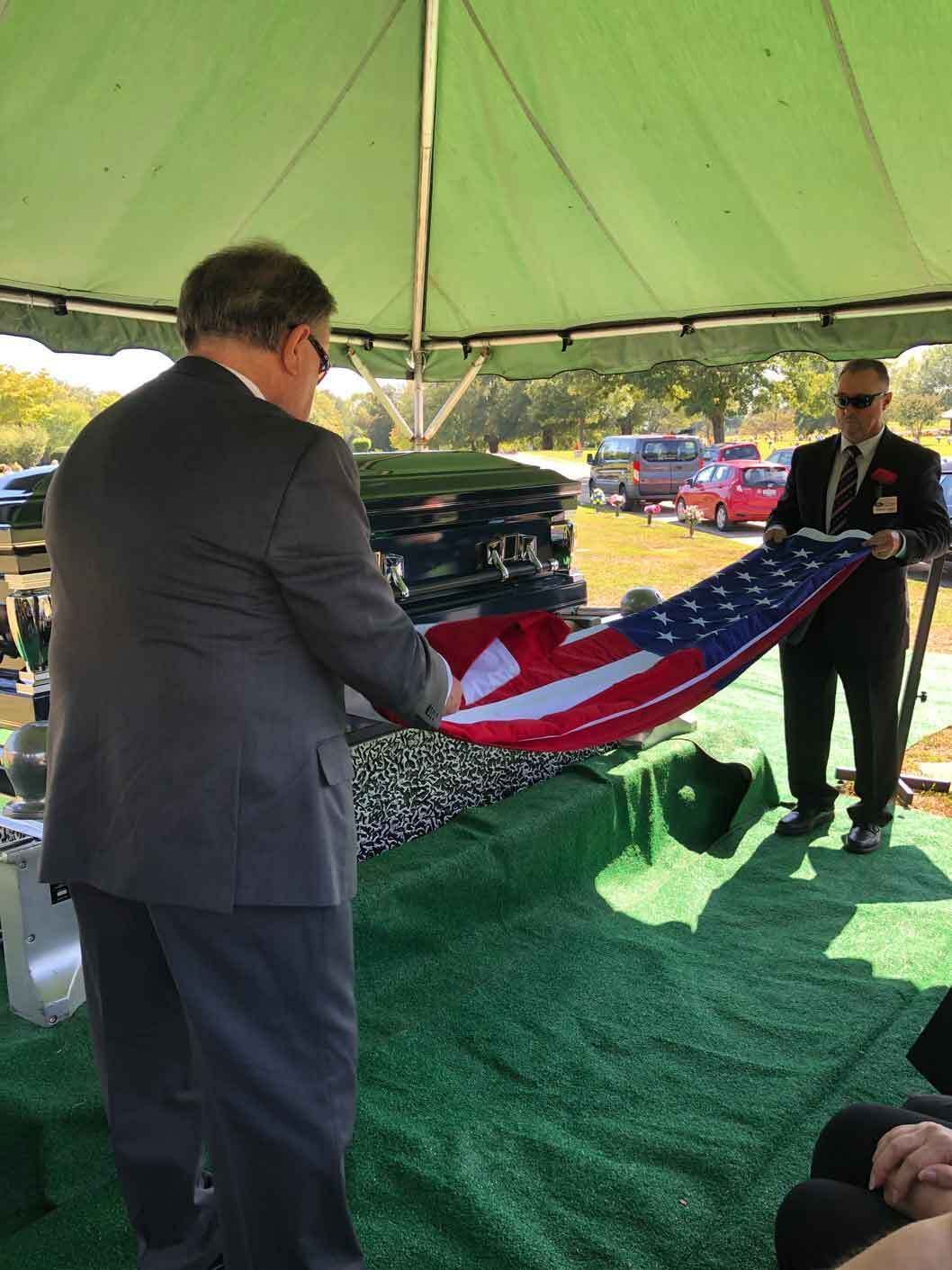Two men folding an American flag over a casket outdoors under a tent.