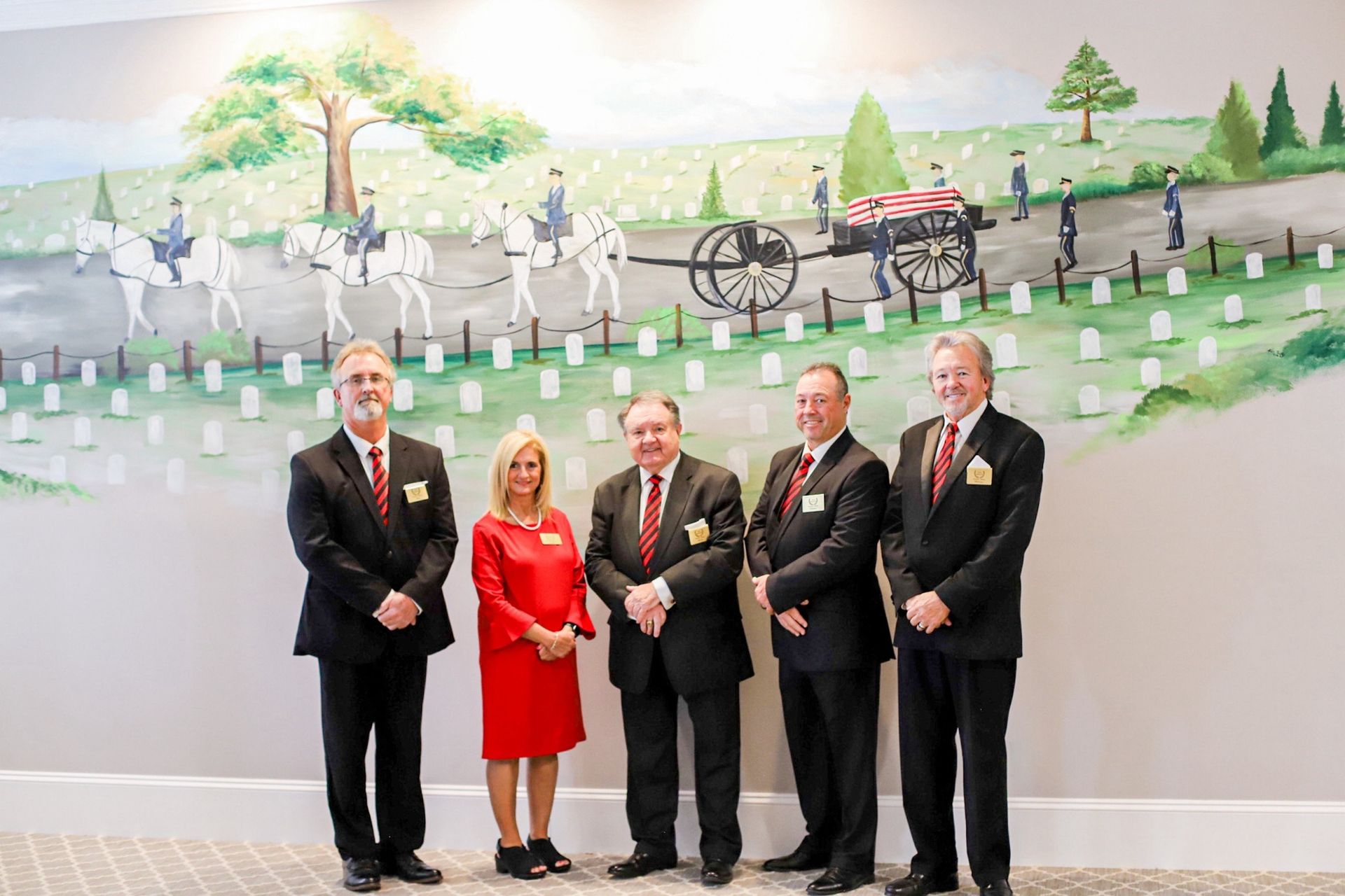 Five people in formal attire stand before a mural depicting a military funeral procession.