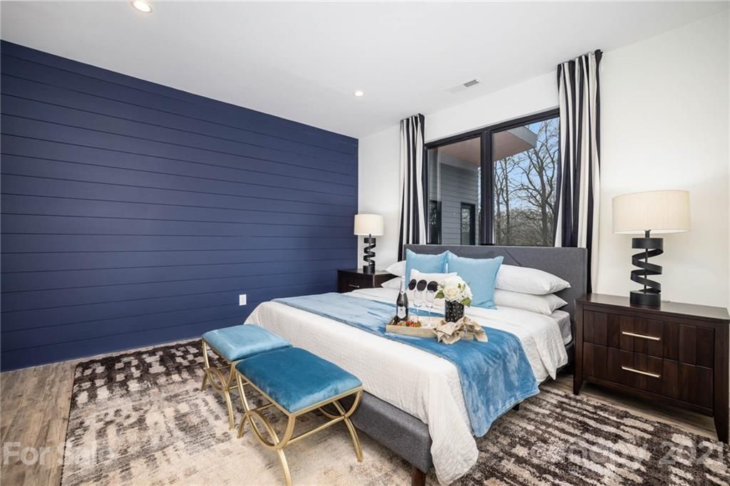 Bedroom with blue accent wall, bed, nightstands, rug, bench, and window with striped curtains.