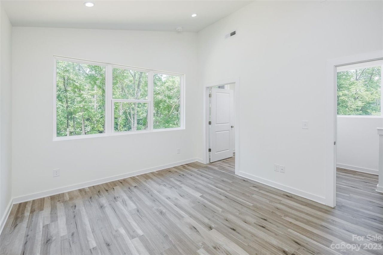 Empty, bright room with gray wood-look floor, large windows, white walls, and doorway.
