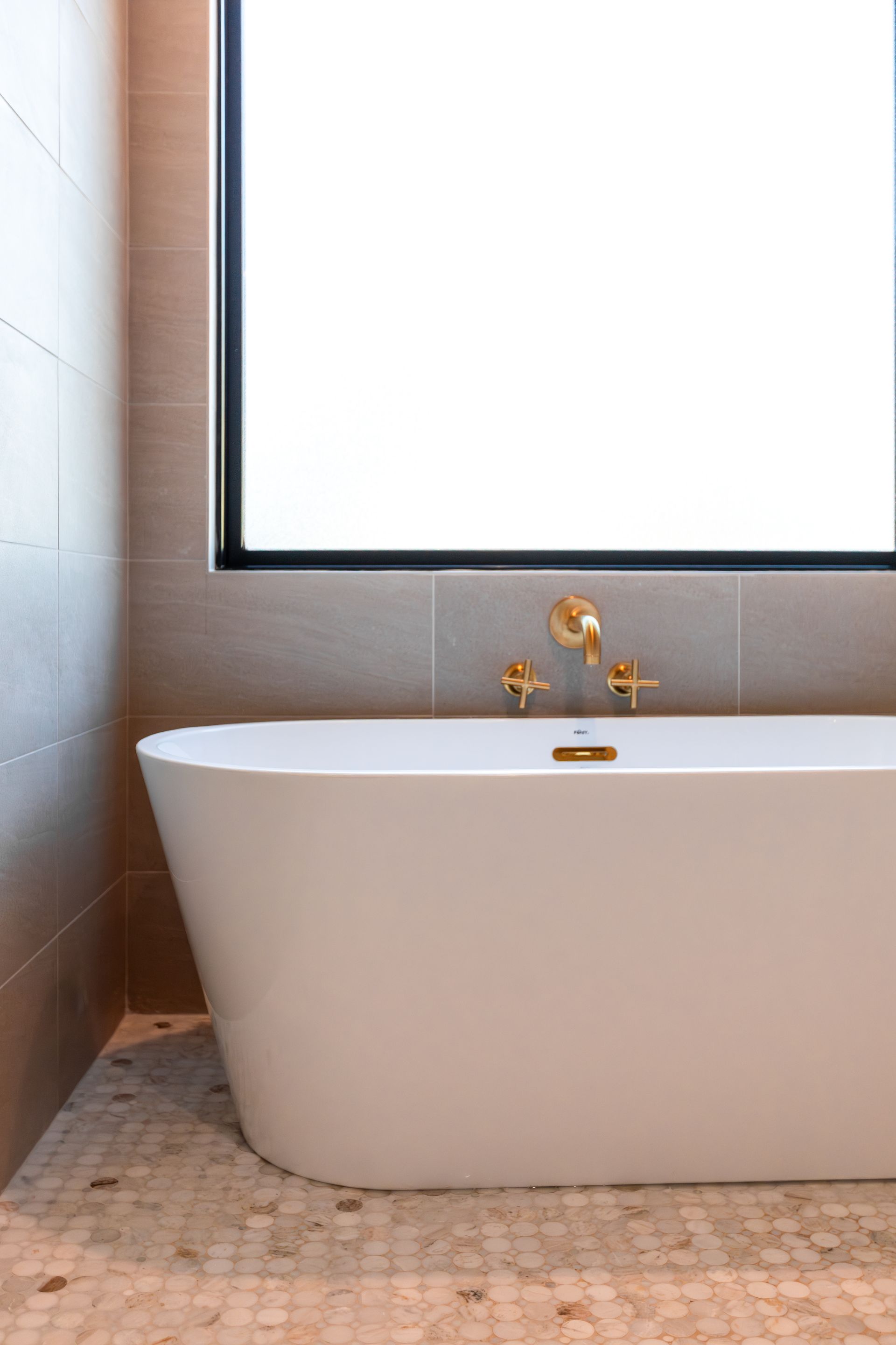 Freestanding white bathtub with gold fixtures near a window in a modern bathroom.