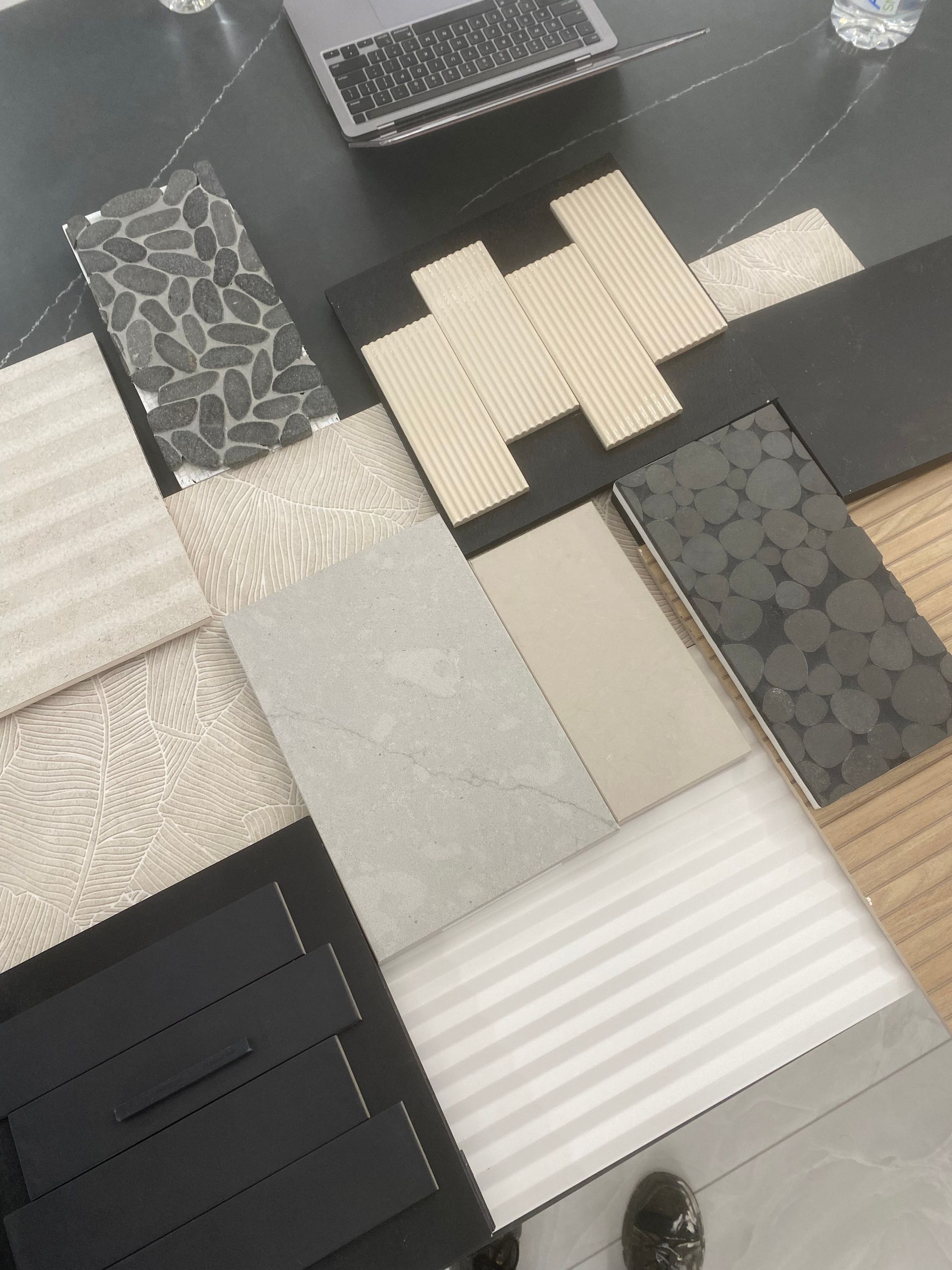 Assorted tile and rug samples on a table. The samples include various textures, patterns, and neutral colors, near a laptop.