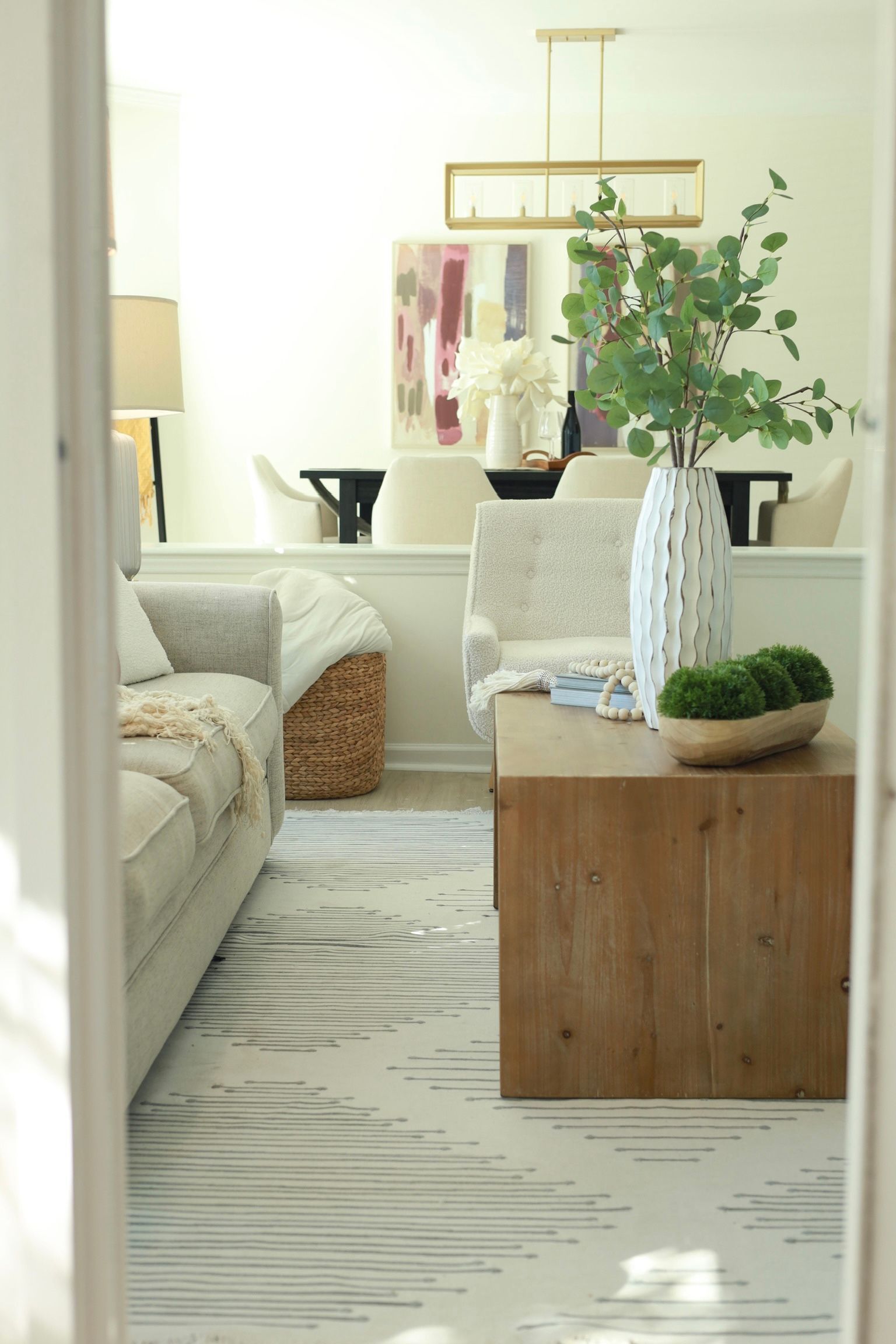 Living room: neutral rug, wooden coffee table, vase with greenery, light-colored sofa, and dining area in background.