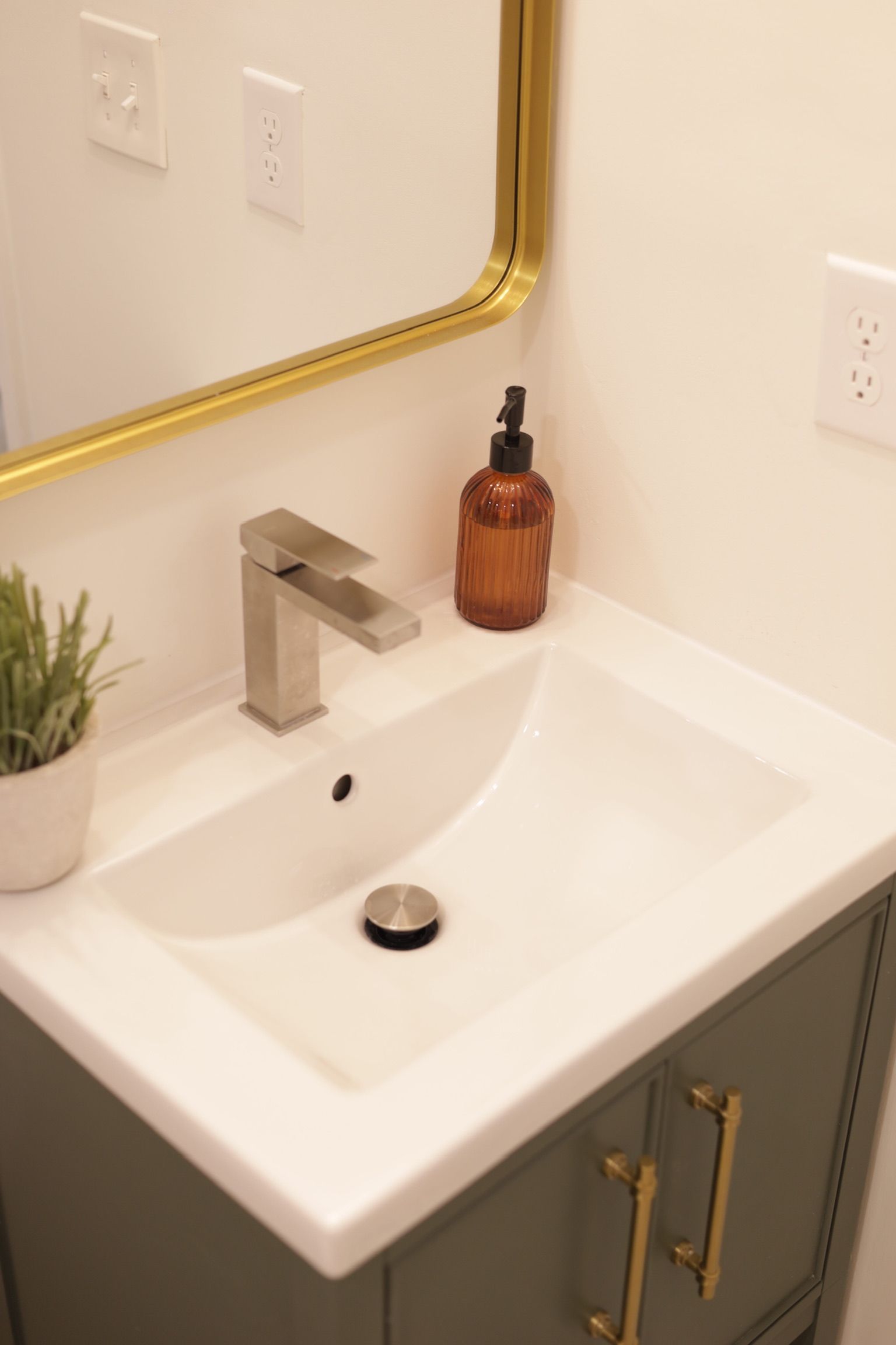 Bathroom sink with gold mirror, grey cabinet, silver faucet, and amber soap dispenser.