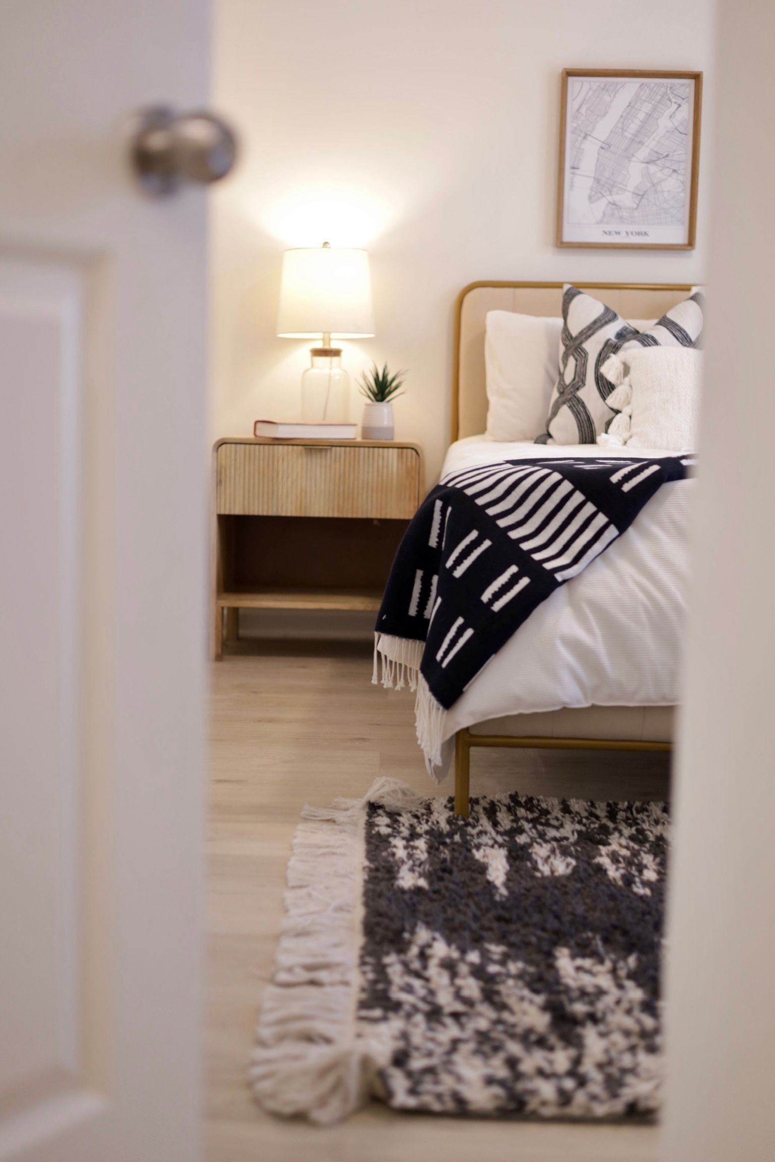 Bedroom interior, viewed through a doorway. Bed with throw blanket, nightstand, rug on the floor.