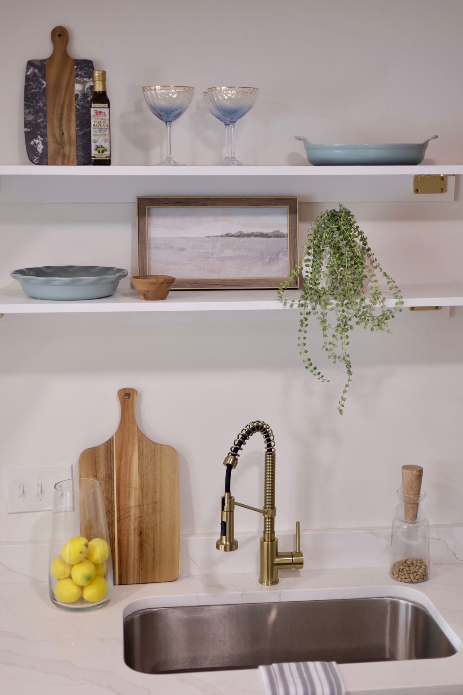 Kitchen with open shelving. Cutting boards, glassware, art, and plant displayed. Gold faucet over sink.