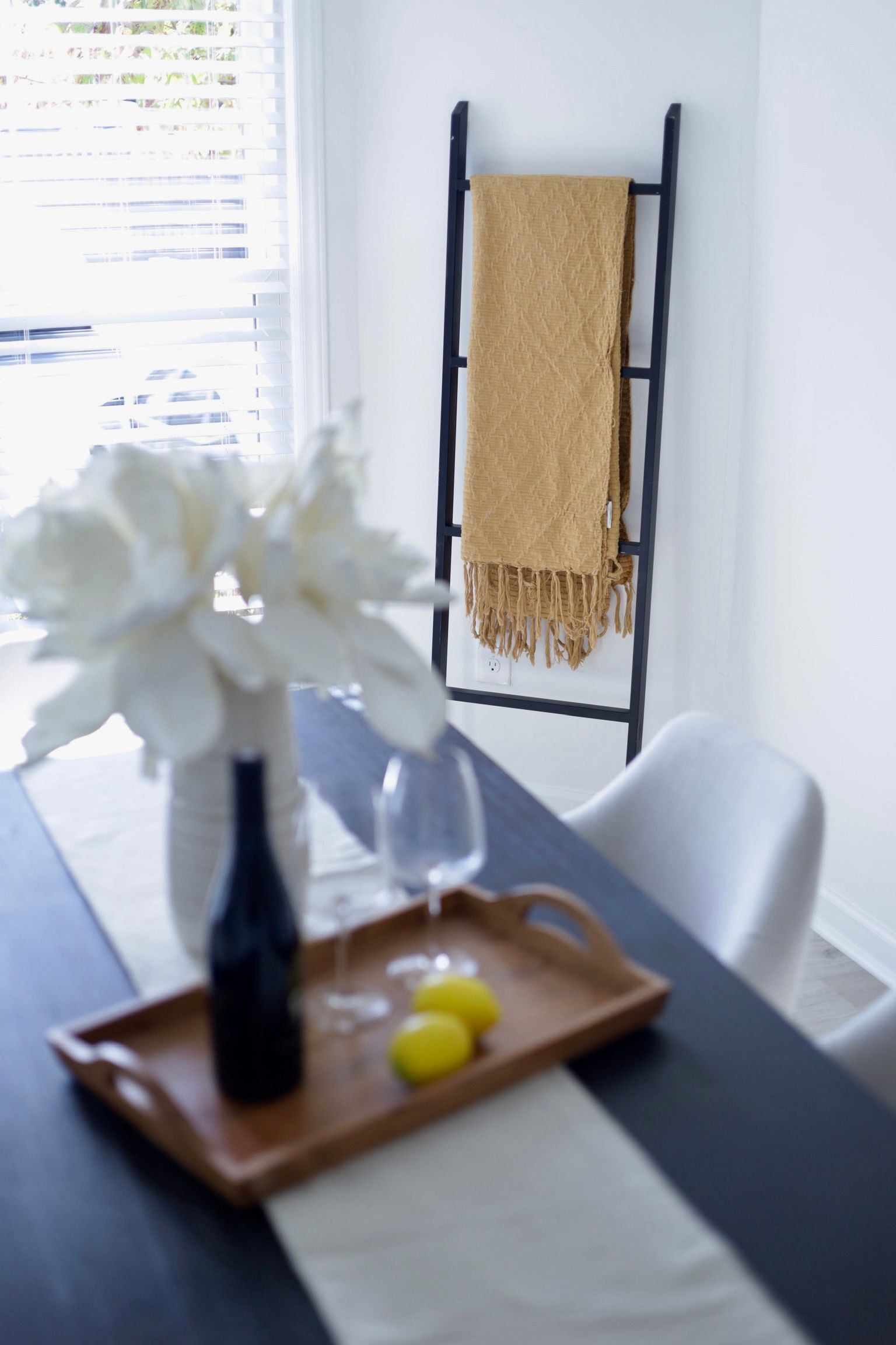 Dining room: Black table with a wooden tray, a ladder with a gold blanket, and a white vase with flowers.