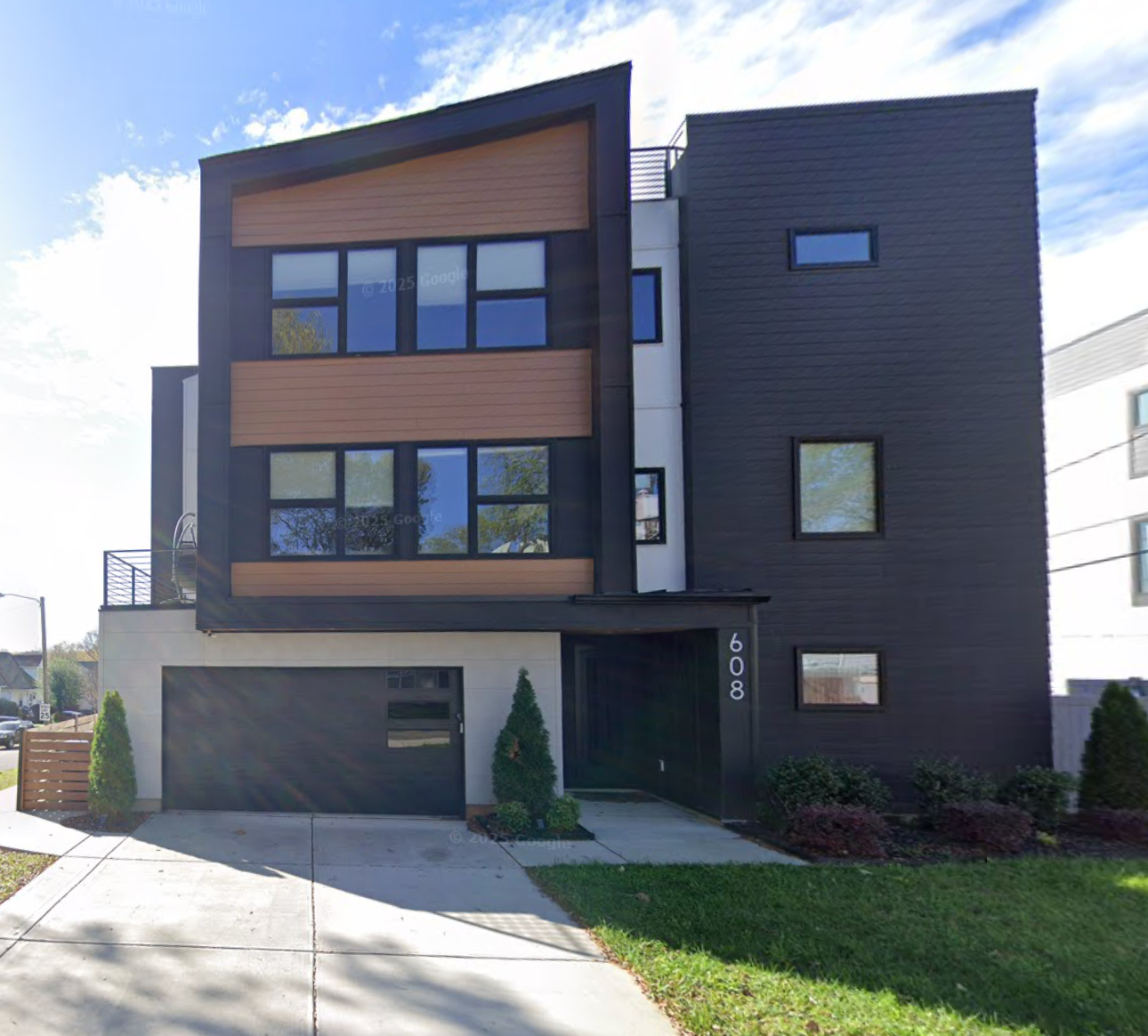 Modern three-story house with dark siding and wood accents; a garage, walkway, and small front yard.