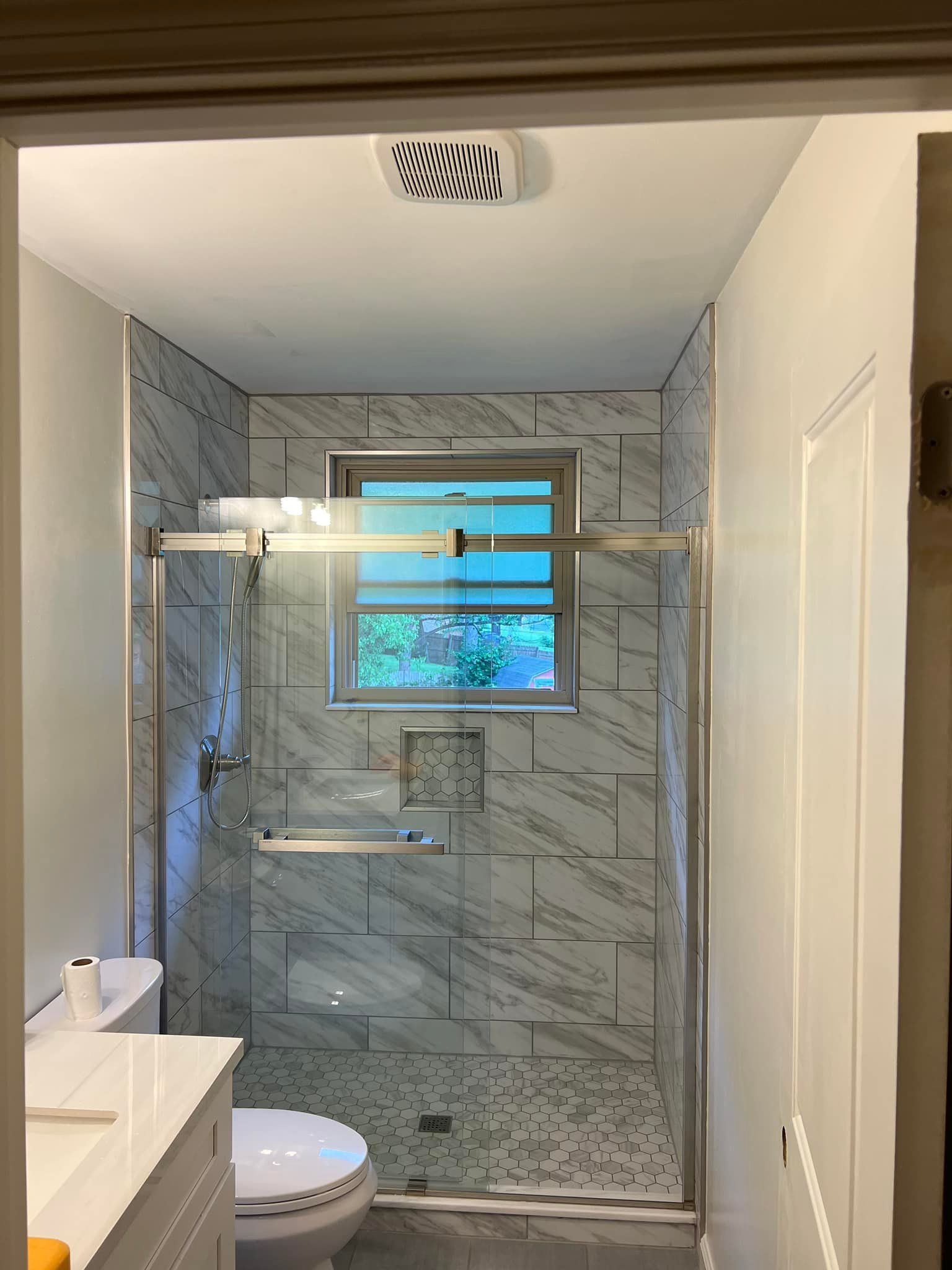 Small bathroom shower with gray tile, glass door, window, toilet, and vanity.