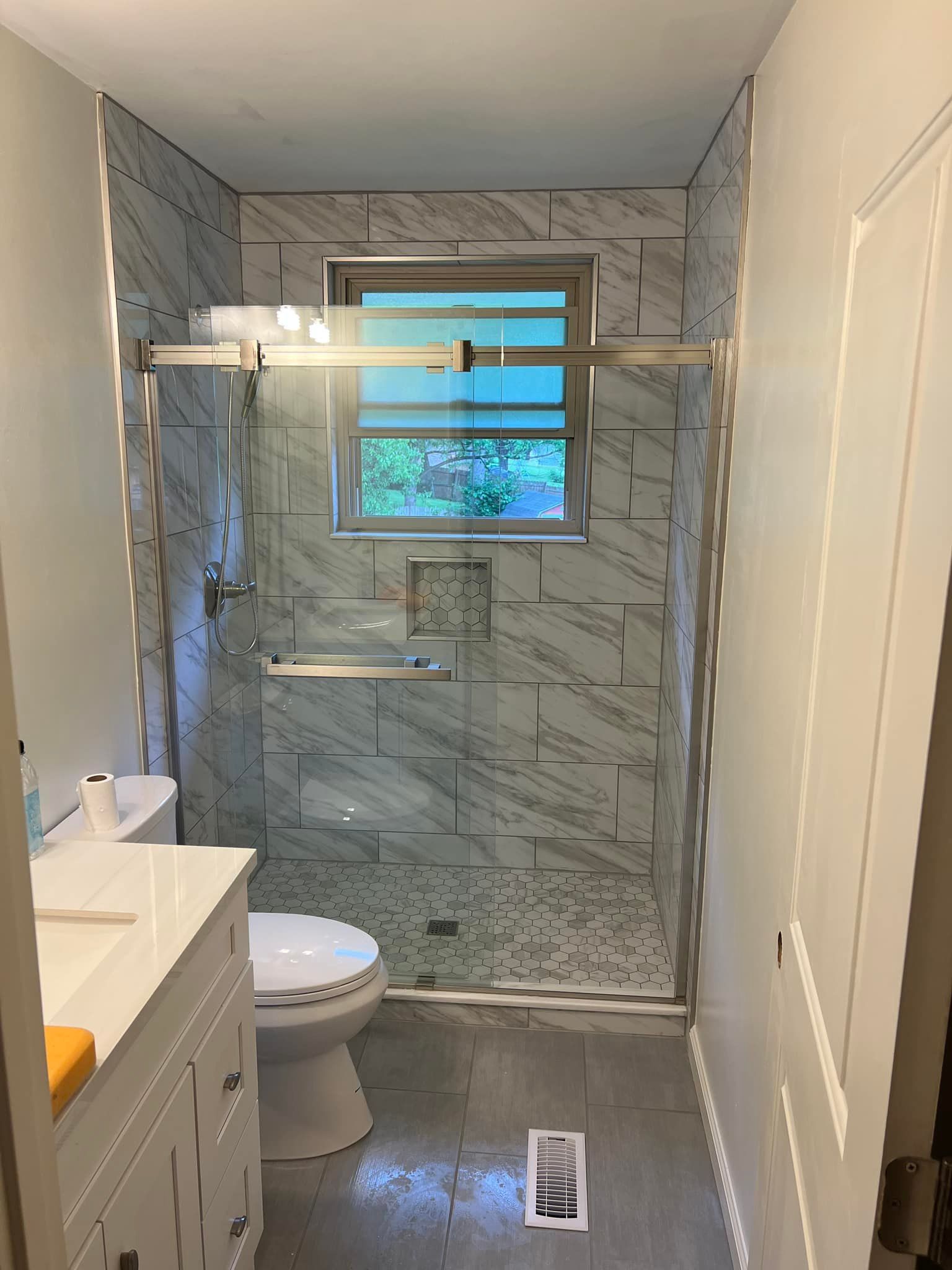 Narrow bathroom with shower, toilet, vanity, and window above a tiled shower wall.