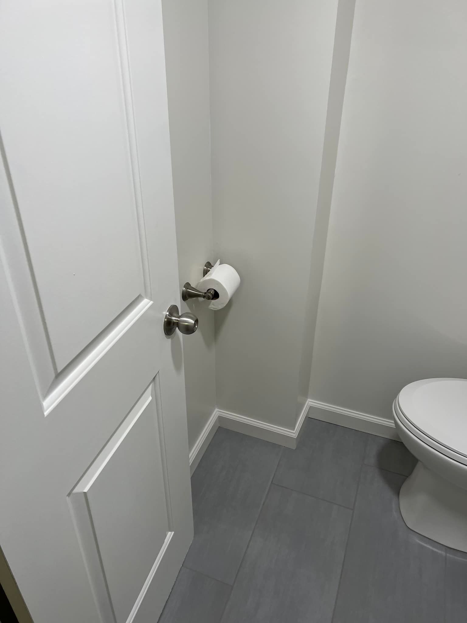 Small bathroom with white door, toilet paper holder, gray tile floor, and partial toilet visible.