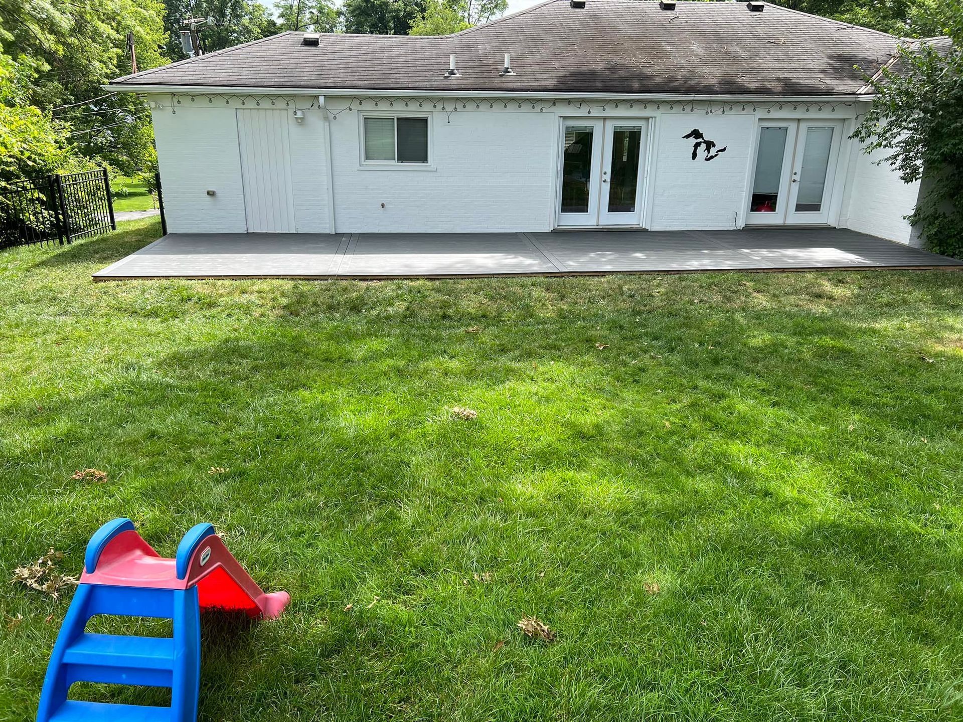 Backyard with white house and patio, green lawn, and a red-and-blue child’s slide in the foreground