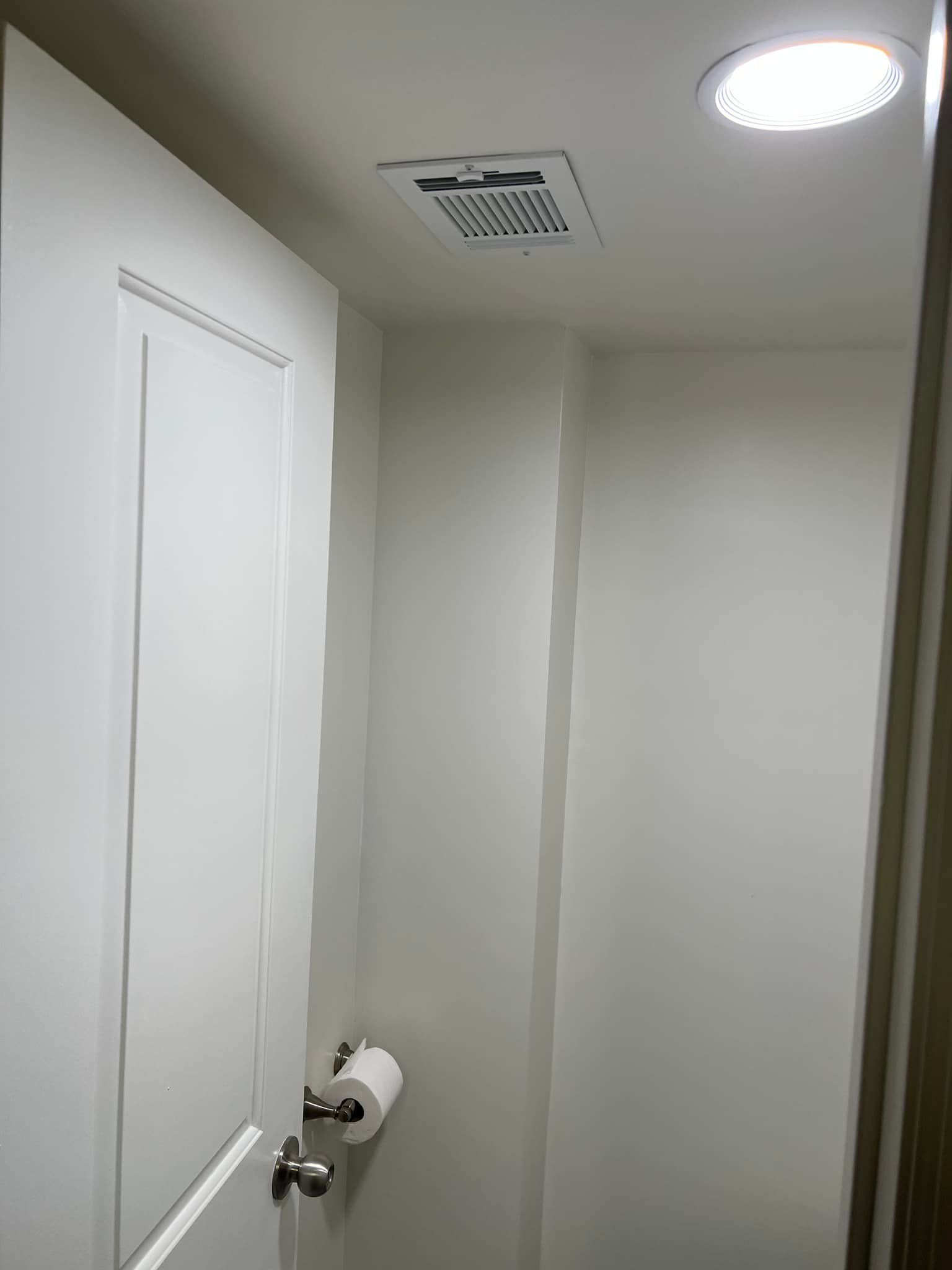Small white bathroom with closed door, toilet paper holder, vent, and ceiling light.
