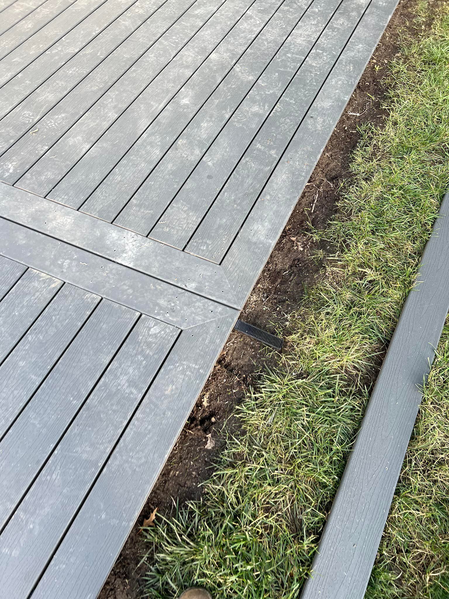 Gray wooden deck beside a narrow strip of soil and grass next to a paved edge