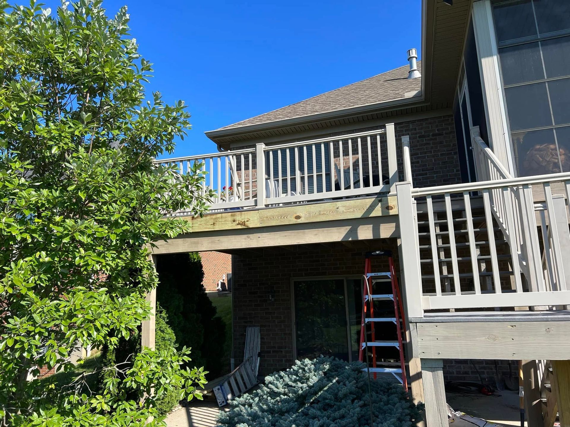 Backyard deck with white railings, ladder, and leafy tree beside a house under a blue sky