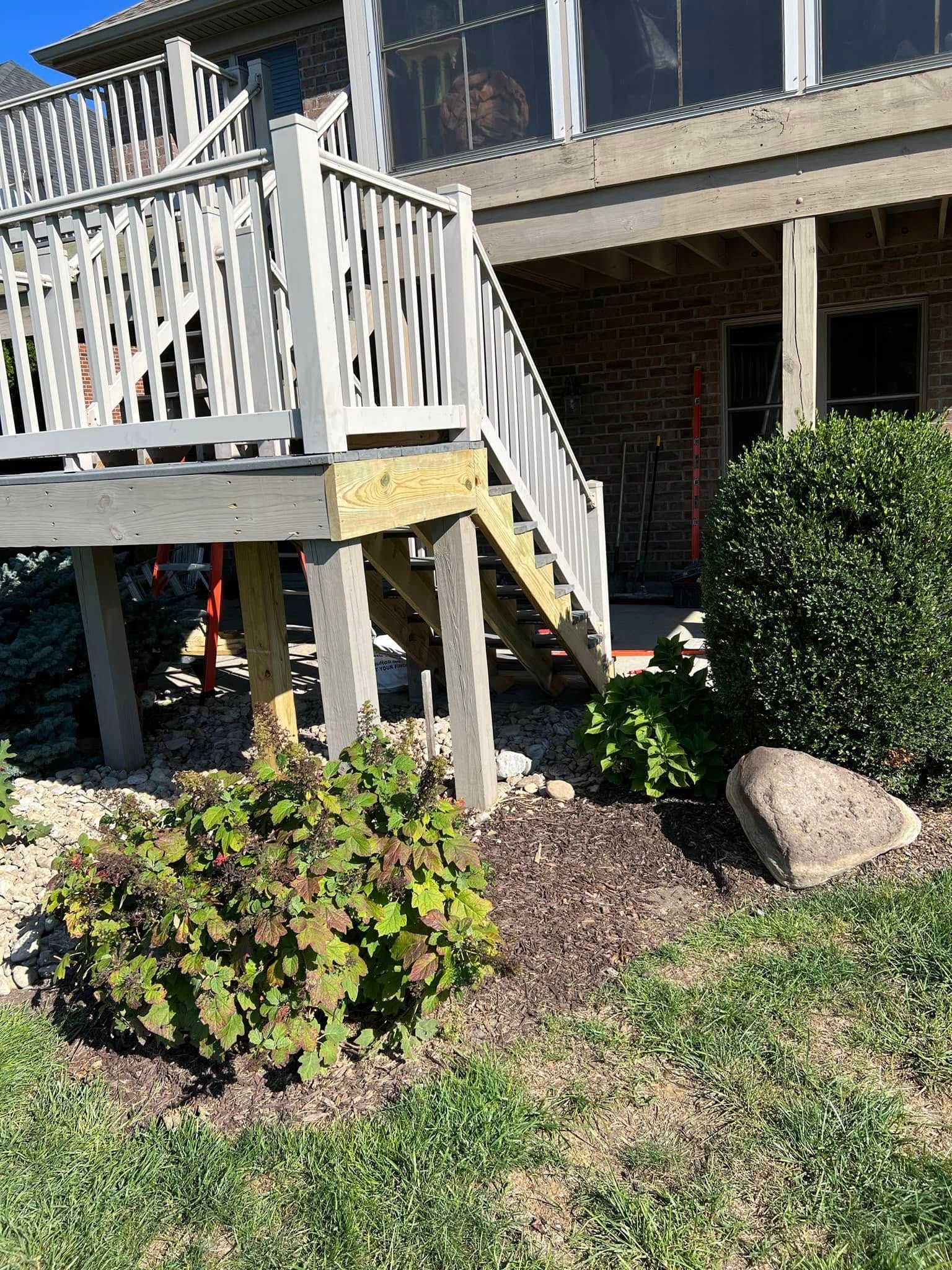 Wooden staircase and porch beside a brick house, with shrubs and landscaping rocks in front.