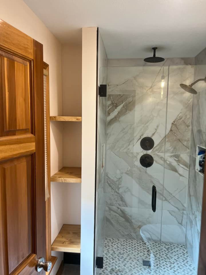 Bathroom shower with glass door, marble-look walls, and wooden shelving beside a wooden door