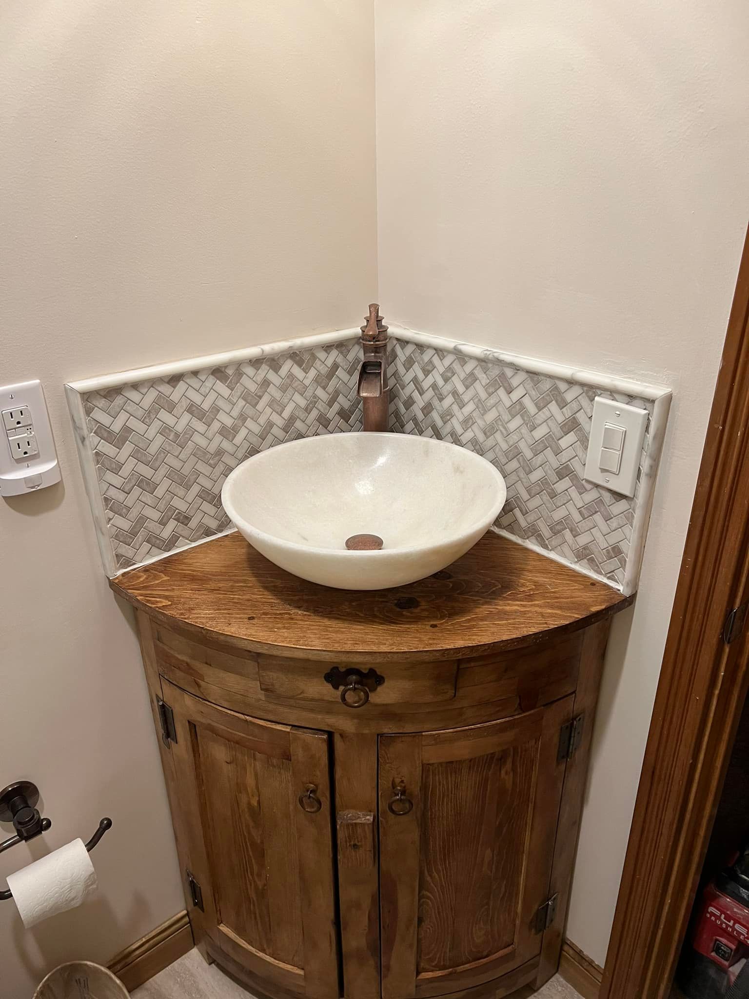 Corner bathroom vanity with wood cabinet, white vessel sink, and patterned tile backsplash.
