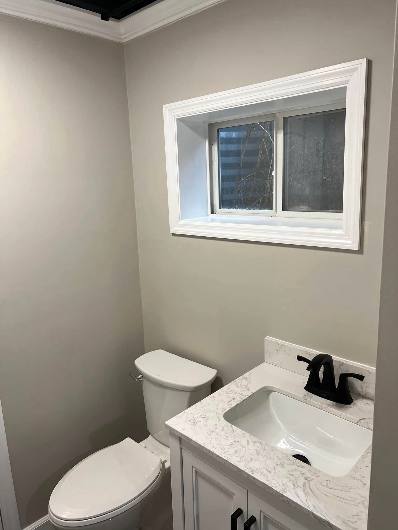 Small bathroom with toilet, white vanity sink, gray walls, and a window above the sink.