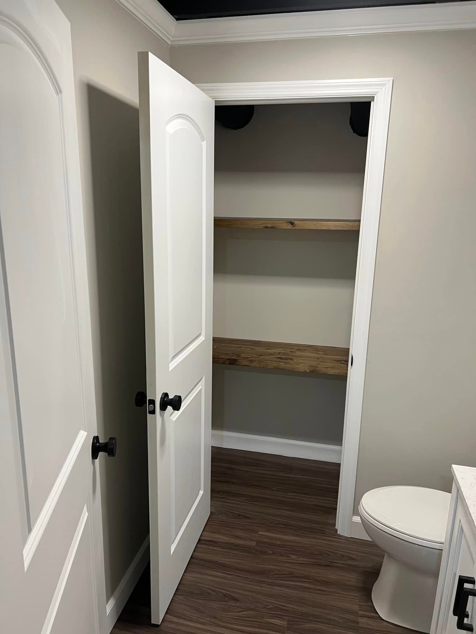 Open bathroom door revealing a closet-like alcove with shelves, beside a toilet and white walls.