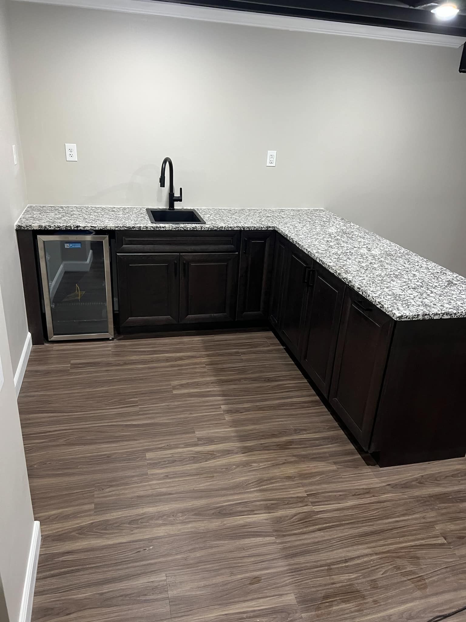 Empty kitchenette with dark cabinets, speckled countertop, sink, and mini fridge against a gray wall.