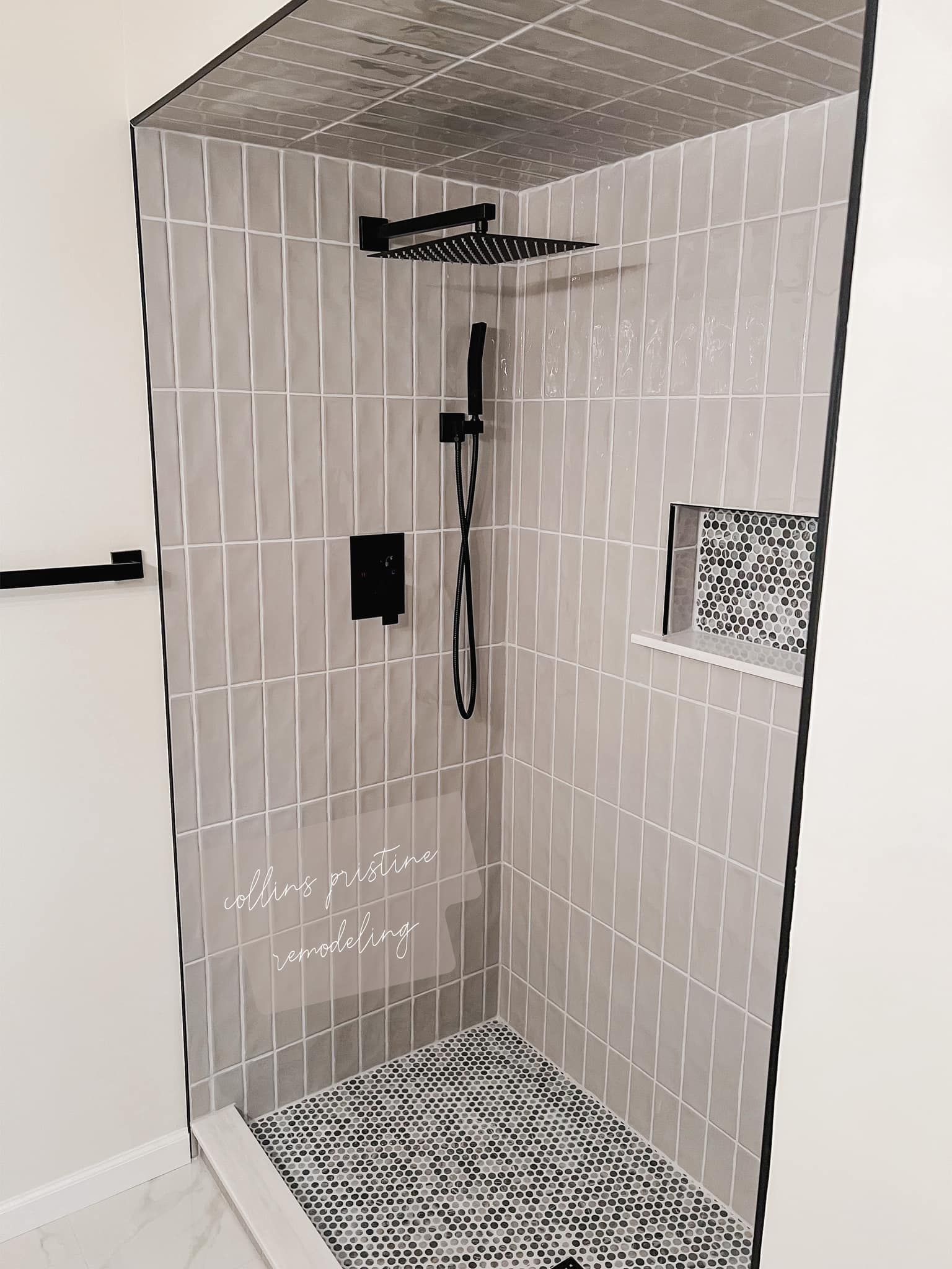 Small tiled shower stall with a black showerhead, wall control, and patterned floor mat.
