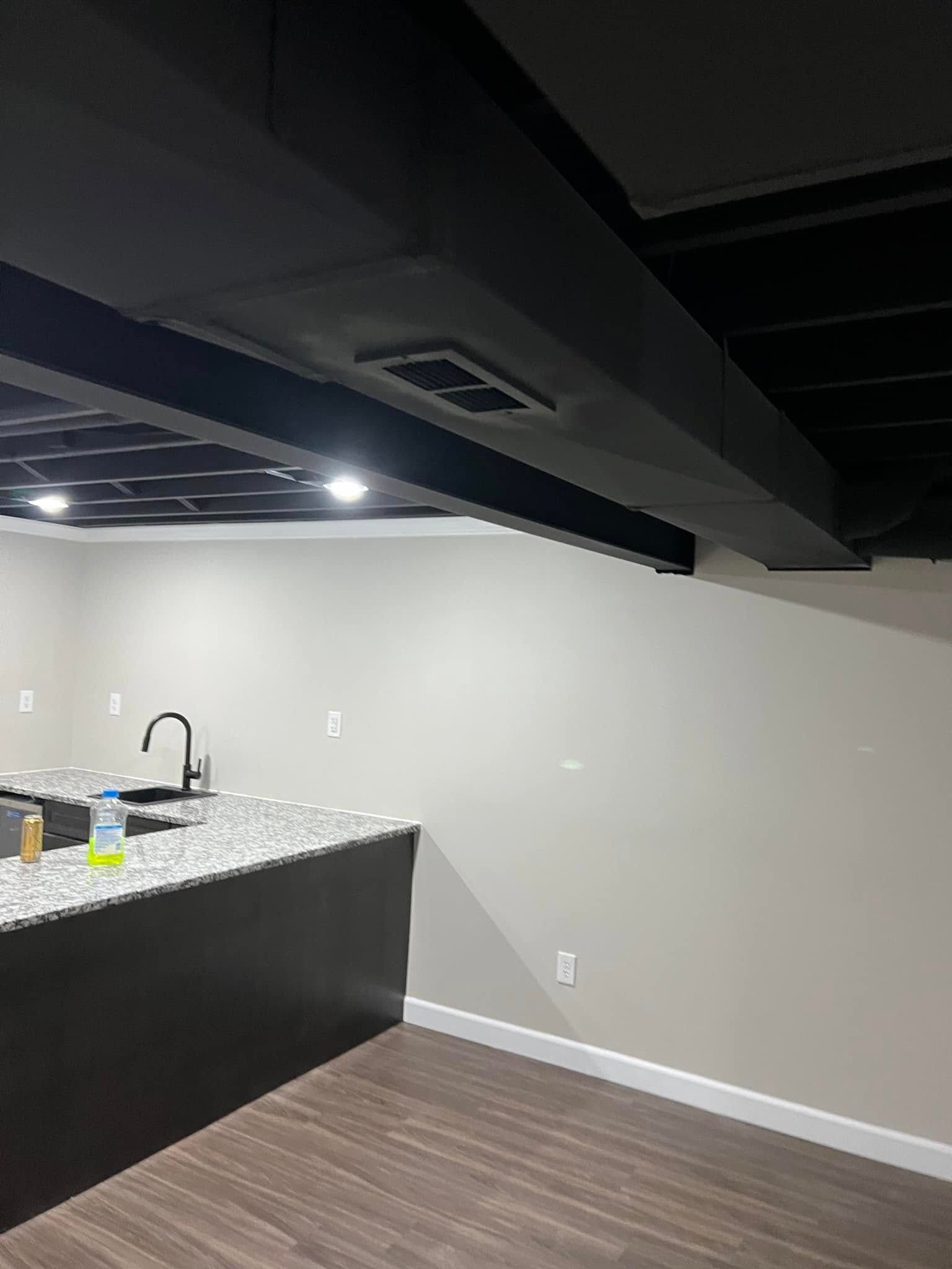 Empty modern basement kitchenette with black ceiling beams, gray walls, and speckled countertop.