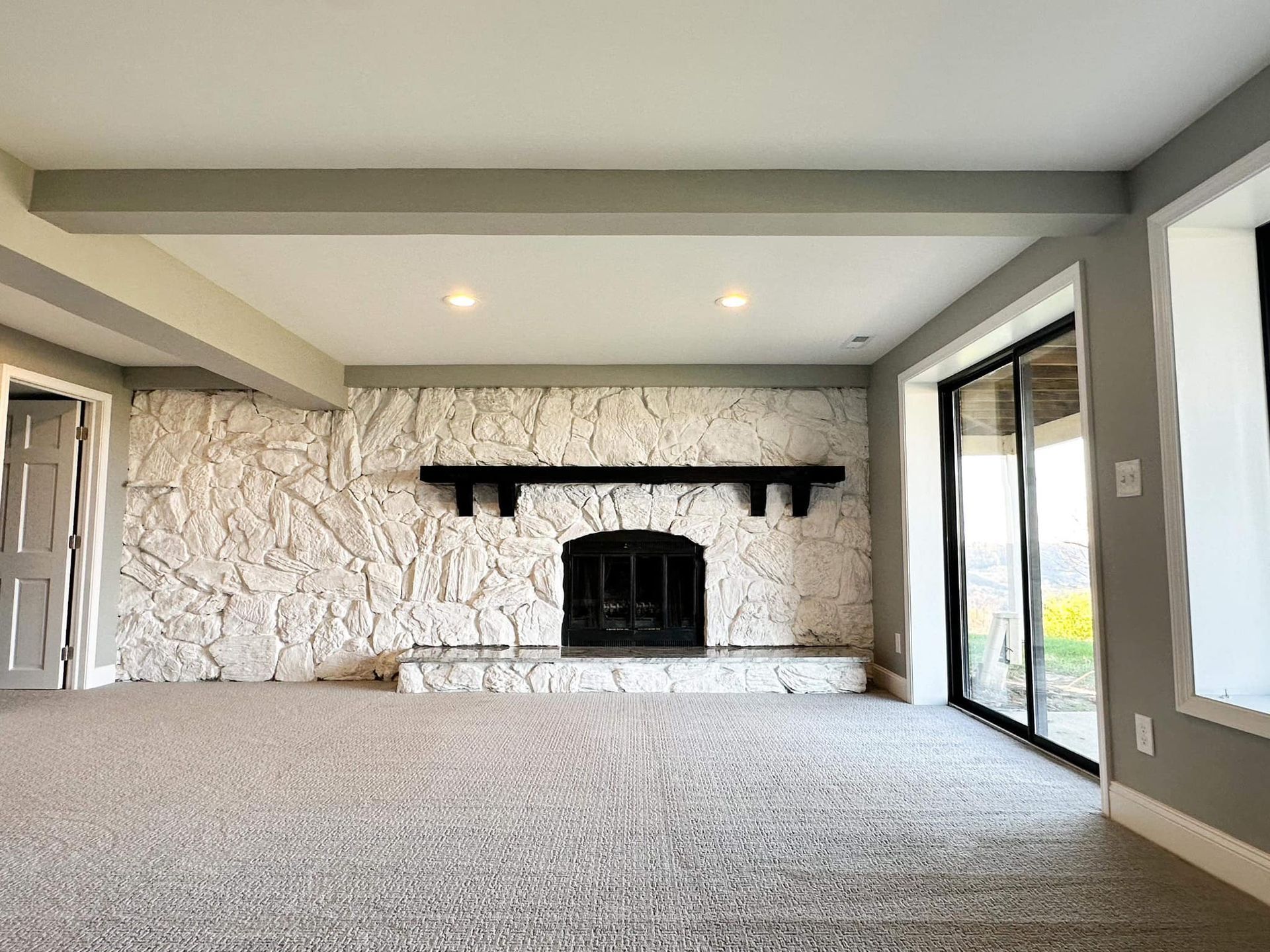 Empty living room with stone fireplace, gray carpet, and large windows overlooking greenery
