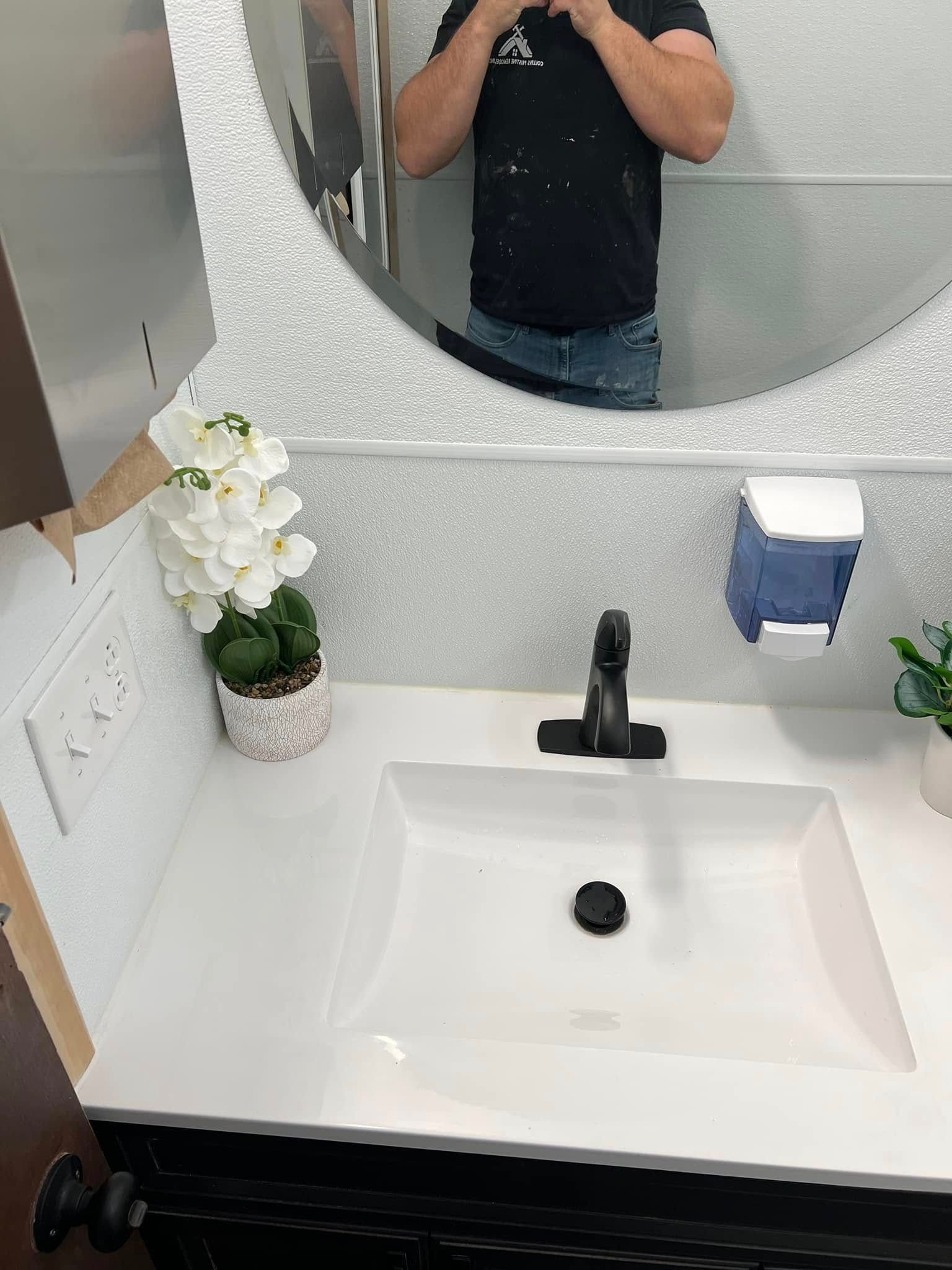 Small white bathroom sink with black faucet, round mirror, and flowers on the counter
