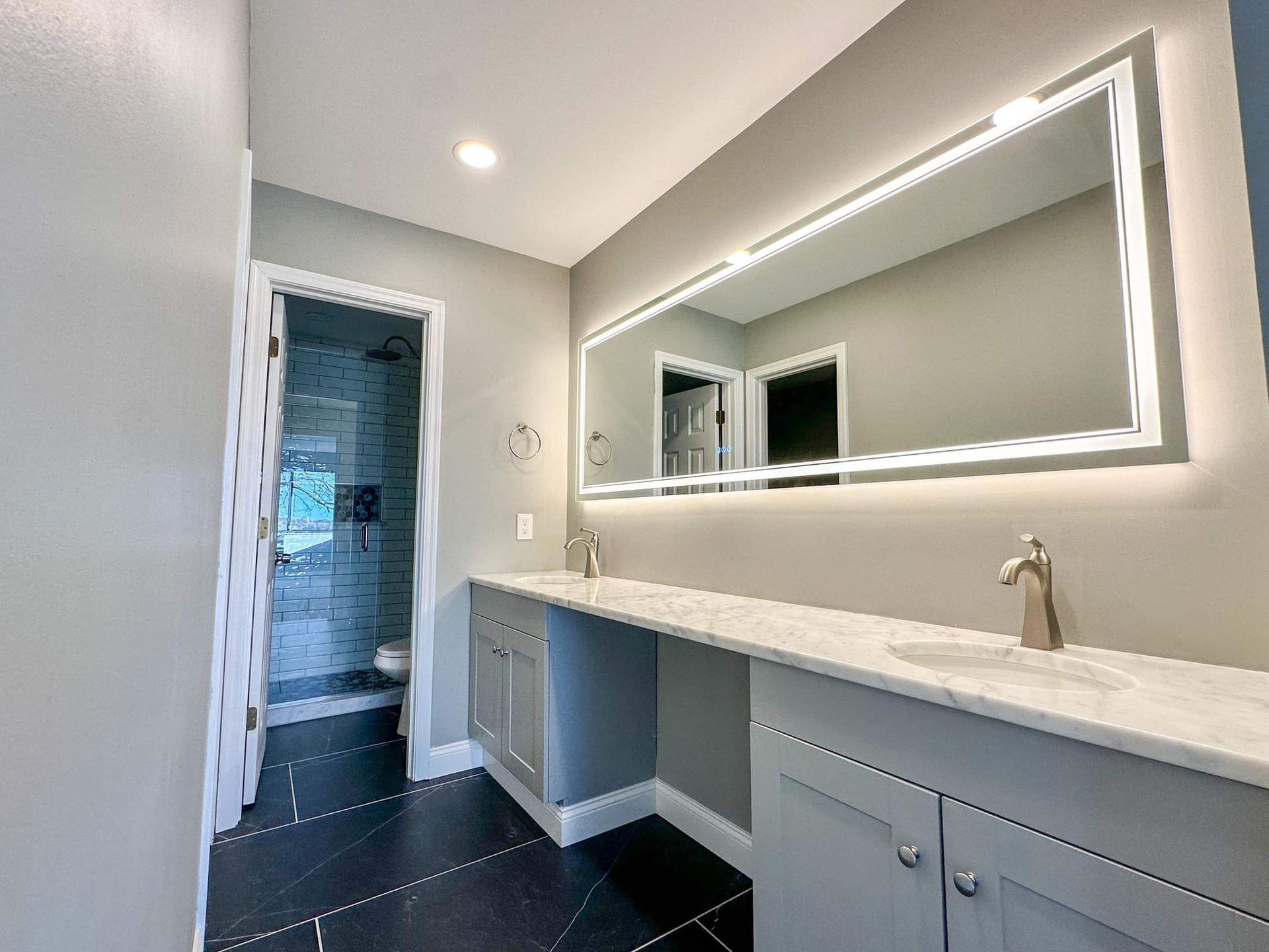 Modern bathroom vanity with large lit mirror, white countertop, and open doorway to a shower area