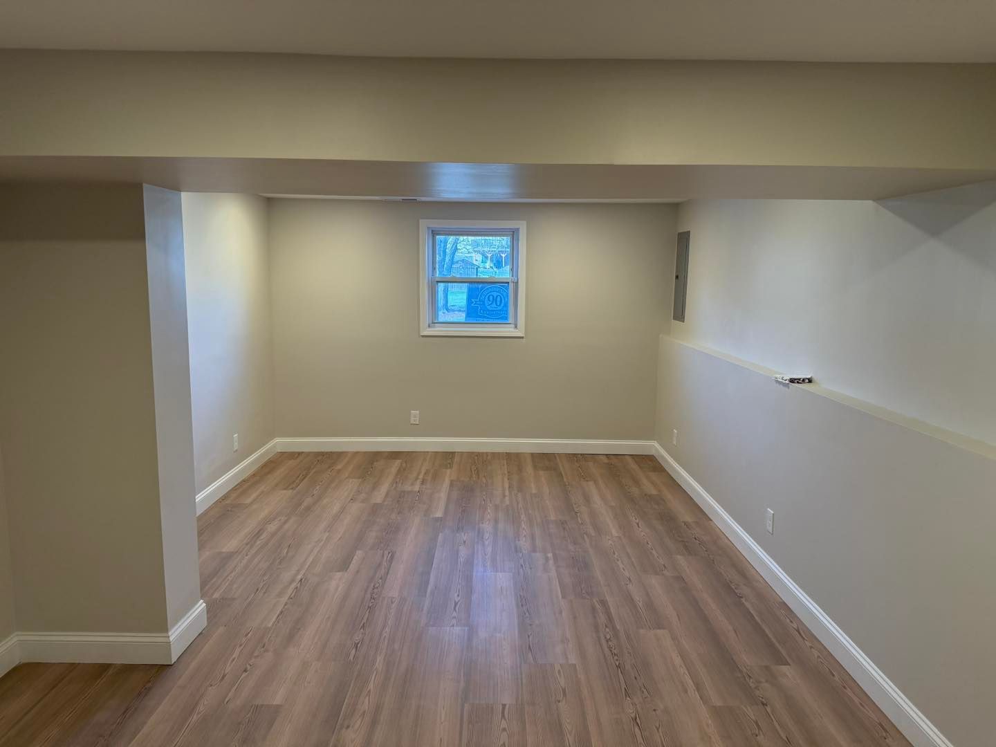 Empty finished basement room with wood flooring, beige walls, and a small window.