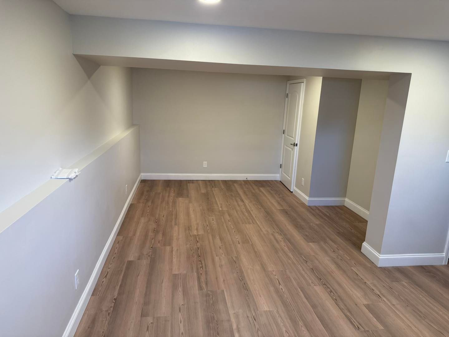 Empty beige room with wood-look flooring and a white door in the back corner.