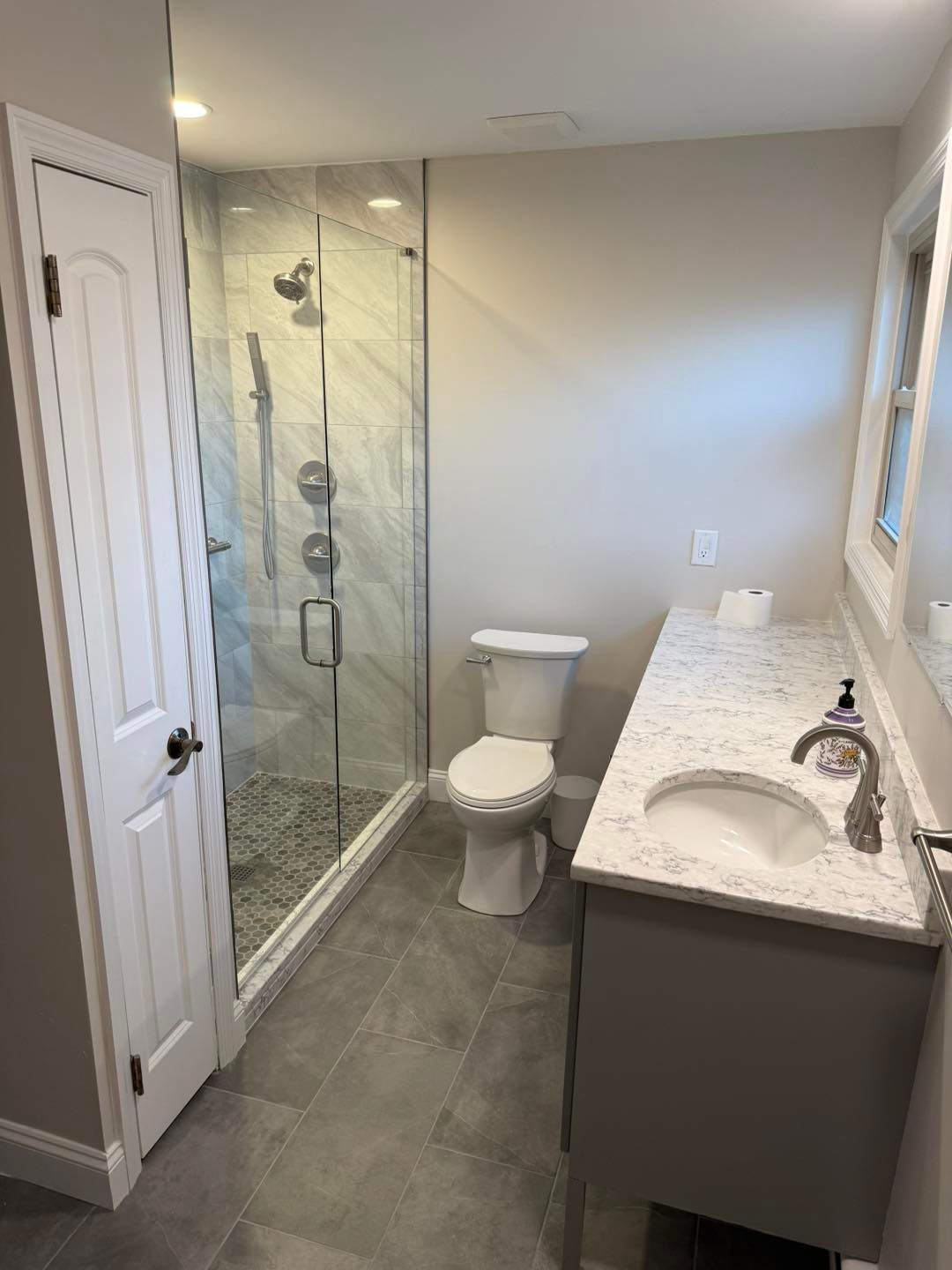 Small bathroom with glass shower, toilet, vanity, and gray tile flooring.