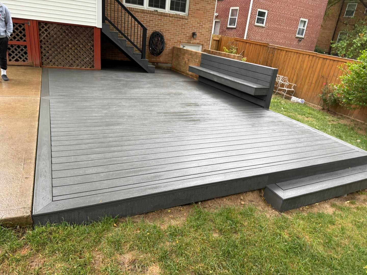 Raised gray wooden deck in a backyard beside brick houses, with steps and a bench-like railing.