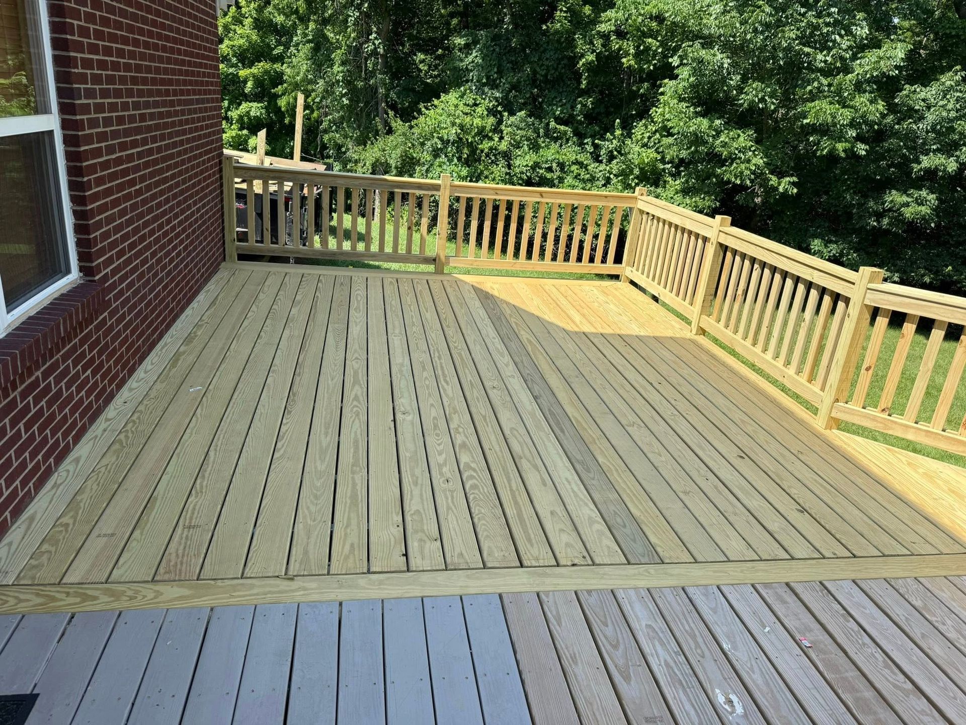 Wooden backyard deck with tan railings beside a brick house, overlooking green trees.