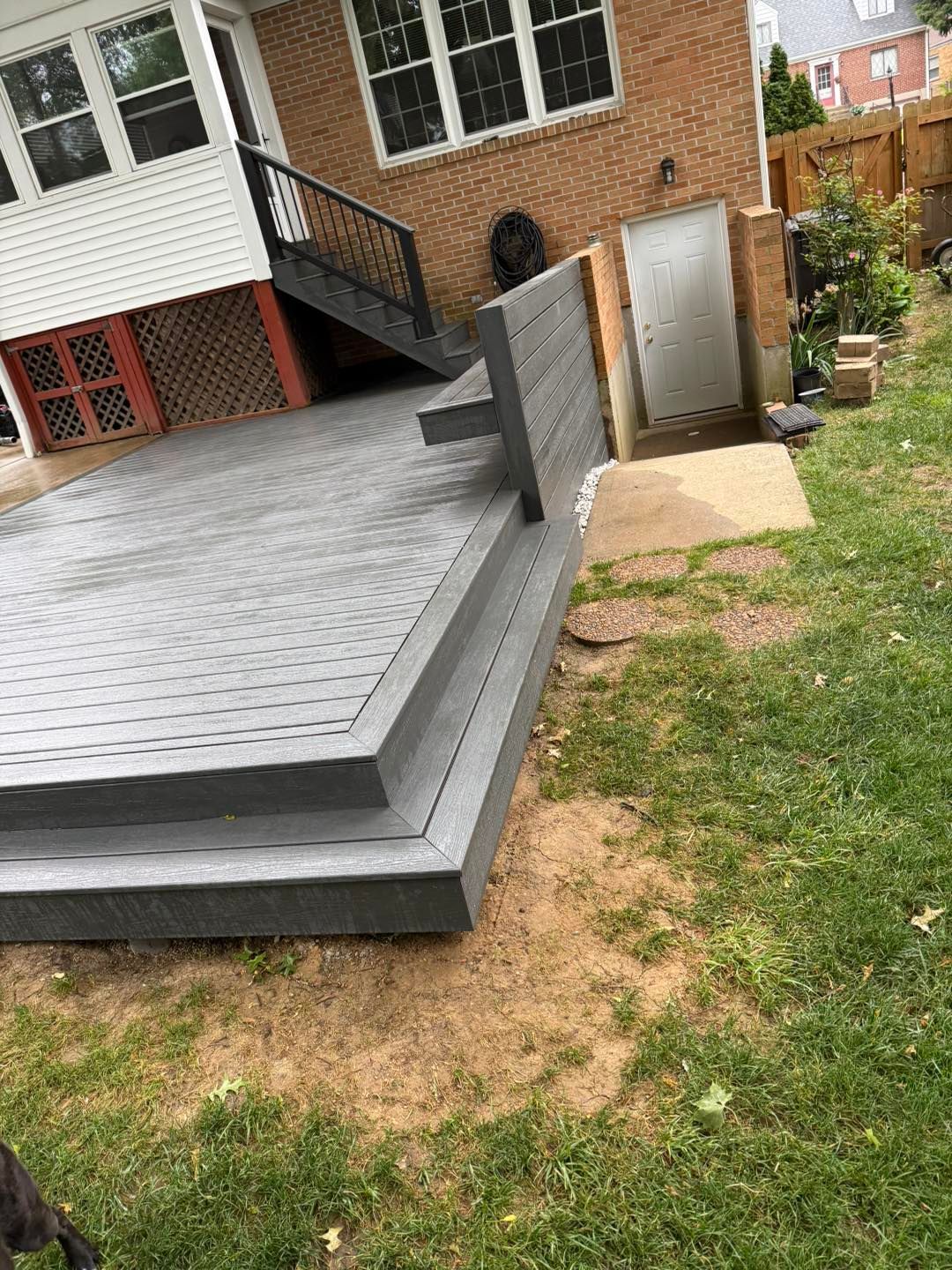 Backyard deck with gray railing, steps, and a small concrete entry beside a brick house.