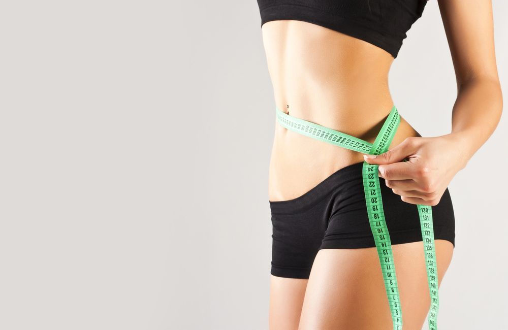 Woman In Black Shorts And Top Measures Waist With Green Tape Against A Gray Background — Brisbane Upper GI Surgery in Chermside, QLD