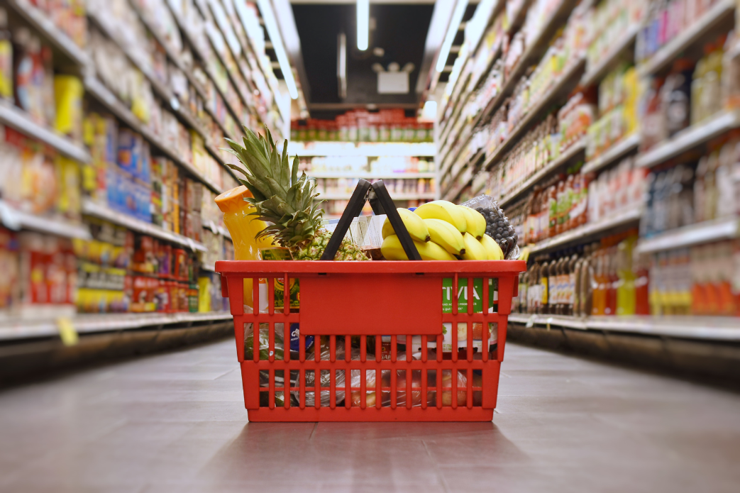 Red shopping basket with groceries in a supermarket aisle, including bananas and a pineapple.