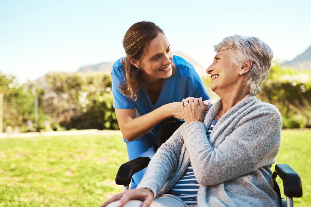 Caregiver smiling and talking with an older woman outdoors in a wheelchair on a sunny day