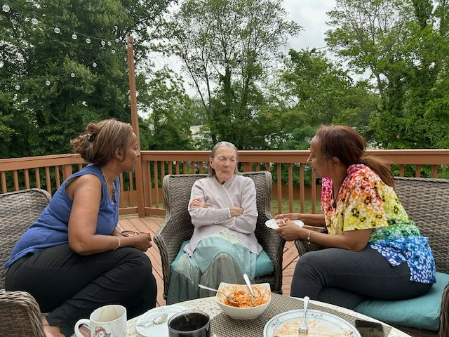 Three people seated on a deck talking, with food on the table.