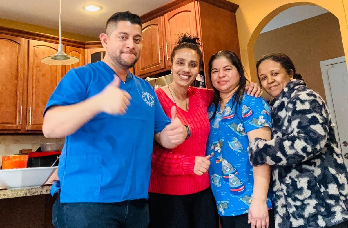 Four people smile and pose for a photo in a kitchen; two people in the front give a thumbs-up gesture.