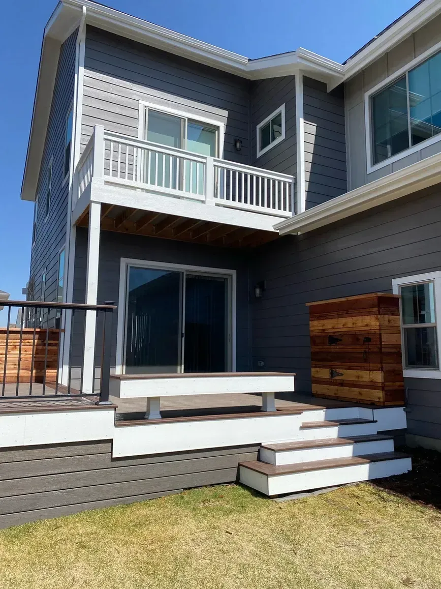 Back of a two-story gray house with deck and stairs leading down to a grassy yard on a sunny day.