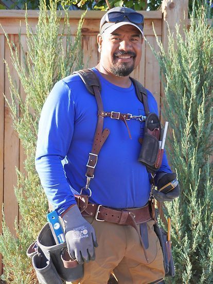 Man in blue shirt and tool belt smiles, standing in front of a wooden fence and greenery.