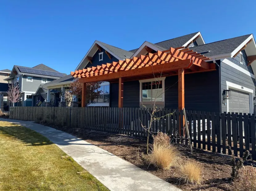 Dark gray house with wooden pergola over the porch, black picket fence, and sidewalk on a sunny day.